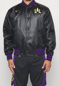 Chaqueta bomber de satén negra con puños y dobladillo de canalé a rayas moradas, que presenta "LAL" bordado en amarillo en el pecho y el logo de Nike en amarillo.