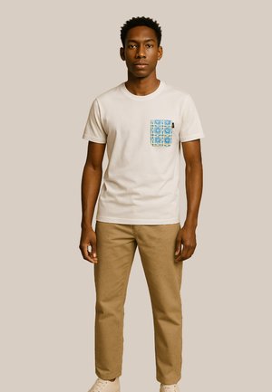 T-shirt crème à manches courtes avec une poche à motifs bleus, associée à un pantalon kaki et des chaussures claires, présentée sur un fond neutre.