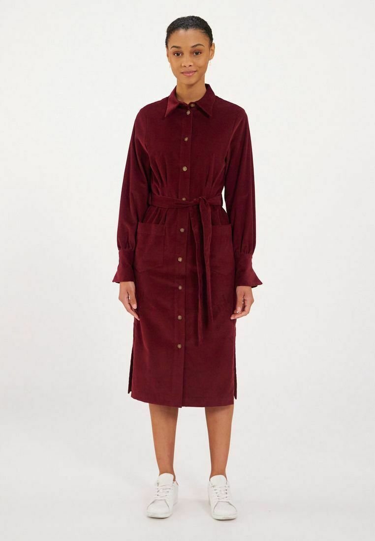 Robe chemise en velours côtelé bordeaux avec un devant à boutons, taille à nouer, manches longues et fentes sur les côtés, associée à des baskets blanches.