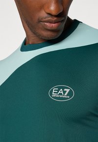 Camisa atlética verde con detalles en azul claro, con textura de malla y cuello redondo. Logo "EA7 Emporio Armani" en blanco.