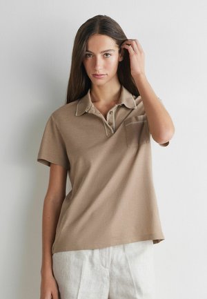 Kvinde med langt brunt hår iført en beige kortærmet poloshirt og lyse bukser, rører ved sit hår og står foran en ensfarvet baggrund.