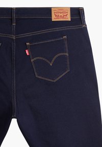 Jean bleu foncé en denim avec une couture contrastante jaune, présentant une poche arrière avec un design en forme de V signature et une étiquette en cuir.