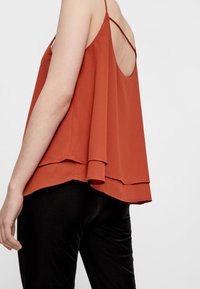 Rdzawopomarańczowa, bezrękawnikowa bluzka o warstwowej konstrukcji i niskim plecach; uszyta z miękkiego materiału, z cienkimi ramiączkami i swobodnym krojem.