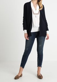 Granatowy kardigan na białej bluzce z koronkowymi akcentami, w połączeniu z dopasowanymi dżinsami i balerinami w panterkę. Codzienny strój o zróżnicowanych fakturach.