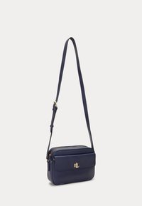 Tmavě modrá kožená kabelka přes rameno s nastavitelným popruhem, zapínáním na zip, přední kapsou a zlatým logem. Hladká textura a obdélníkový tvar.