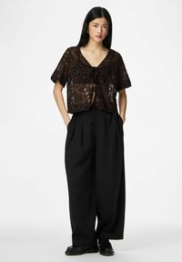 Kortermet, brun blondebluse med blomsterprint, sløyfe i halsen, kombinert med høytlivs svarte vide bukser og svarte sko.