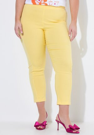 Persoon die cropped gele broek en felroze sandalen met hoge hakken en grote strikken draagt, staand tegen een witte achtergrond.