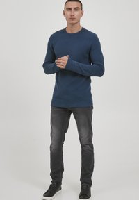 Pleteni pulover s mornarskim prugama, okruglim izrezom i rebrastim manžetama, u kombinaciji s tamno sivim trapericama i crnim niskim tenisicama. Uski kroj.