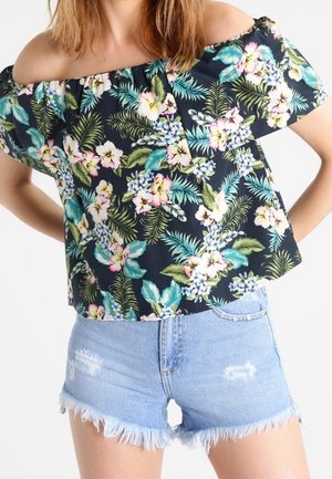 Femme portant un haut noir épaules dénudées avec un imprimé floral tropical coloré et un short en jean bleu clair usé avec des ourlets effilochés.