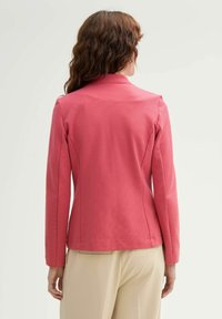 Blazer rose ajusté avec une finition texturée, épaules structurées et manches longues, associé à un pantalon beige clair.