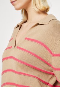Beżowy dzianinowy sweter polo w różowe paski, z dekoltem w kształcie litery V i krótkimi rękawami. Tekstura wydaje się gładka, a fason jest luźny.