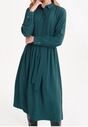 Frau trägt ein langärmeliges, türkisfarbenes Midikleid mit plissiertem Vorderteil, geknöpften Manschetten und einem Taillengürtel.