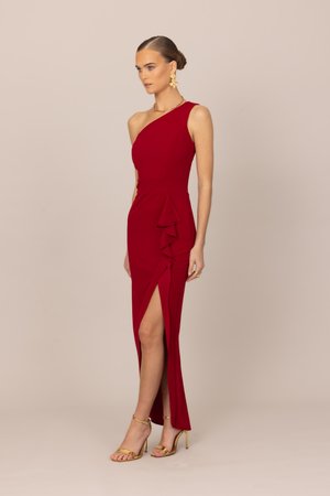 Kvinde i rød aftenkjole med ét skulderstrop, højt sidespalt og flæsedetalje, iført gyldne højhælede sandaler og iøjnefaldende øreringe.