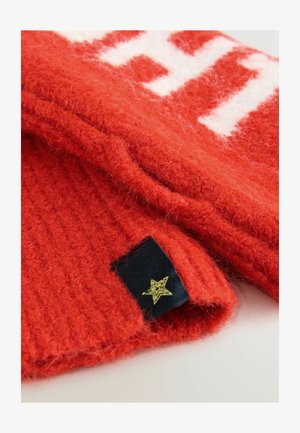 Rød strikket sweater med hvidt mønster, der har ribbet tekstur og en vævet label med en gylden stjernedetalje. Blødt og fuzzet materiale.