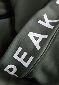 Mörkgrön tyg med en texturerad yta; djärva vita bokstäver delvis synliga, med en sömdetalj längs kanten.