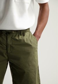 Khaki-Shorts aus leichtem Stoff mit elastischem Bund, Seitentaschen und dezentem Glanz; kombiniert mit einem schlichten weißen T-Shirt.