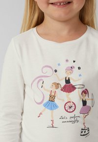 Weißes Langarmhemd mit drei gezeichneten Figuren in bunten Röcken, schwarzen Oberteilen und Accessoires, mit dem Text "Lasst uns erstaunlich performen."