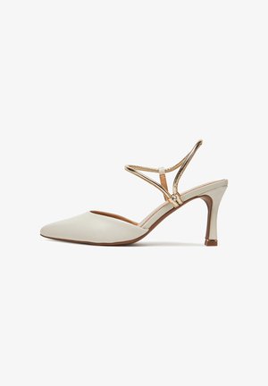 Beige műbőr magassarkú slingback cipő hegyes orral, fémes díszítéssel és keresztben futó pántokkal. Karcsú stiletto sarokkal rendelkezik.