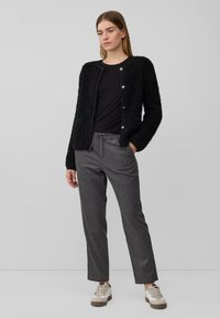 Junge Frau, die einen schwarzen strukturierten Knopf-Cardigan, ein schwarzes Shirt, graue Jogginghosen mit Kordelzug und weiße Sneakers trägt und vor einem schlichten grauen Hintergrund steht.