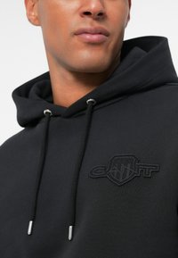 Sudadera negra de tela suave, con un logo bordado en tono sobre el pecho, con cordones y detalles metálicos plateados.