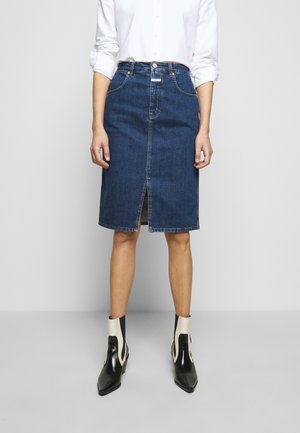 Person wearing a white button-up shirt, knee-length blue denim skirt with front slit, and black and white pointed ankle boots standing against a plain background.