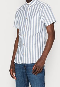 Kortärmad skjorta med knappar i ljusblå och vita vertikala ränder. Har en nedknäppt krage och träknappar. Tillverkad av bomull.