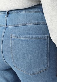Des jeans en denim bleu clair présentent une poche arrière avec des coutures discrètes et une coupe classique. Le matériau a une texture douce avec une légère élasticité.