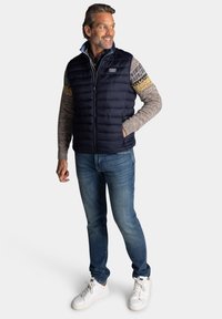Marine blauwe puffer vest over een gebreide trui met patroonmouwen, gecombineerd met blauwe jeans en witte sneakers. Heeft een ritssluiting aan de voorkant en een logo patch.