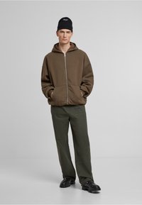 Olivgrüner Reißverschluss-Hoodie mit Fronttaschen, kombiniert mit olivgrünen Hose und schwarzen klobigen Schuhen. Schwarze Strickmütze mit dezentem Logo-Detail.