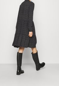 Černé květinové šaty s dlouhými rukávy, zúženým designem a vysokým límcem, spárované s černými koženými botami po kolena. Matný povrch a pevné podrážky.