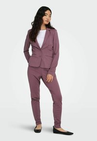 Lilla blazer og matchende bukser laget av et glatt, elastisk stoff. Blazeren har en enkel knapp og noterte slag. Svarte leiligheter fullfører antrekket.