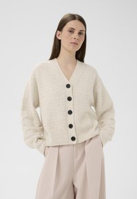 Frau trägt eine strukturierte cremefarbene Strickjacke mit schwarzen Knöpfen und hellbeige hoch taillierte plissierte Hose, steht mit den Händen in den Taschen.