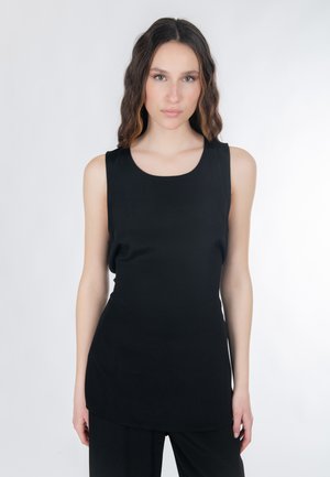 Jeune femme aux longs cheveux ondulés portant un haut noir sans manches et un pantalon noir, debout devant un fond blanc.