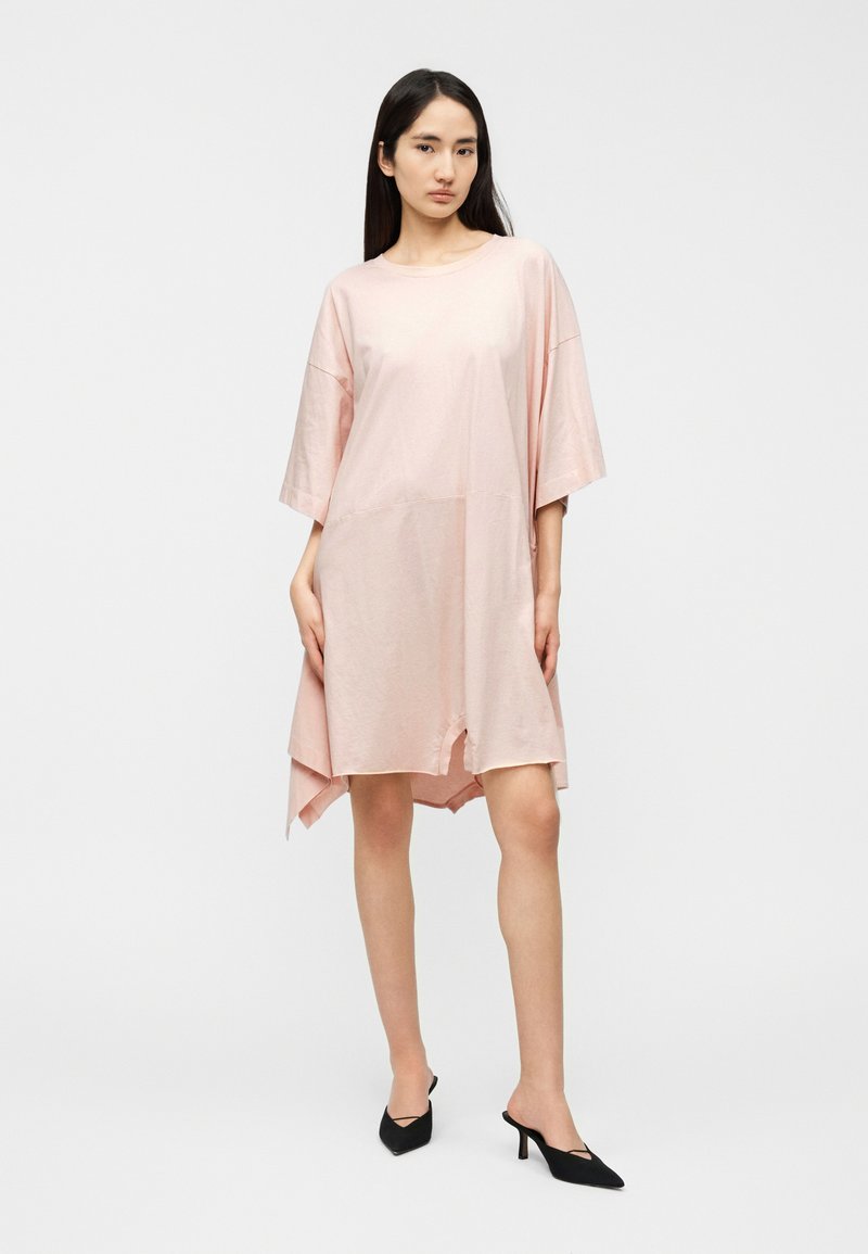 Robe-chemise oversize rose avec manches courtes, encolure arrondie, ourlet à hauteur des genoux, fentes latérales et texture lisse. Stylisée avec des talons noirs.