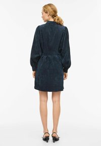 Tamnozelena teksturirana haljina s naduvanim dugim rukavima, pristajućim strukom i ravnom suknjom. Ima ovratnik i manšete sa gumbima.