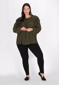 Olivengrøn bluse med rynkede ærmer, strukturerede prikker og rund halsudskæring. Sammen med sorte skinny jeans og flade sko.