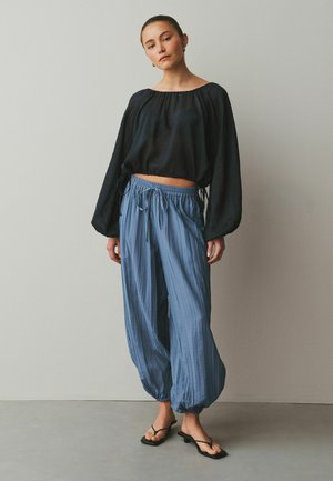 Eine Frau steht vor einer schlichten Wand und trägt ein schwarzes, locker sitzendes, langärmliges Cropped-Top, hellblaue plissierte Balloon-Hosen und schwarze Sandalen mit Absatz.