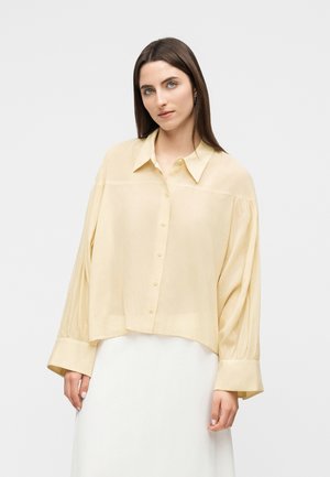 Frau mit glattem braunem Haar, die eine lockere, blassgelbe Bluse mit Knöpfen und einen weißen Rock vor einem einfarbigen Hintergrund trägt.