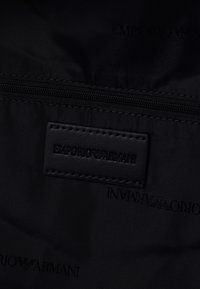 Svart nylon tyg med svart sydd Emporio Armani läderetikett och en svart dragkedja ovanför.