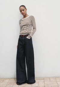 Gestreiftes langärmliges Oberteil in Creme und Braun, kombiniert mit weit geschnittenen dunkelblauen Jeans, die einen braunen Gürtel und auffällige Nähdetails aufweisen.