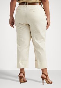 Henkilö, joka käyttää kermahohtoisia cropped-housuja, joissa on napitettu takataskut, sekä ruskeita korkokenkiä, yksinkertaista taustaa vasten.