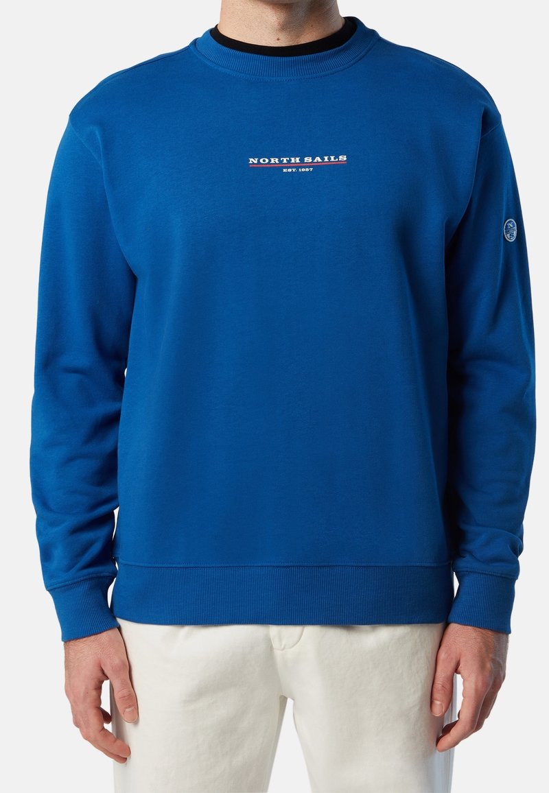 Mann trägt ein blaues Sweatshirt mit Rundhalsausschnitt und dem Schriftzug "North Sails EST. 1957" auf der Brust sowie eine weiße Hose, Hände entspannt an den Seiten.