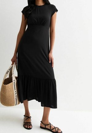 Zwart midi-jurk met kapmouwen, gather aan de taille en een uitlopende, gerimpelde zoom. Combinatie met een geweven tas en zwarte sandaaltjes met banden.