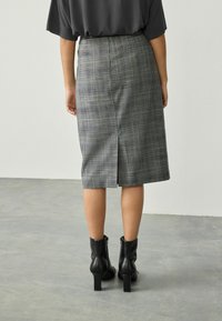 Frau in einem grauen karierten knielangen Rock mit einem Schlitz hinten und schwarzen hochhackigen Stiefeletten, die auf einem grauen Boden steht.