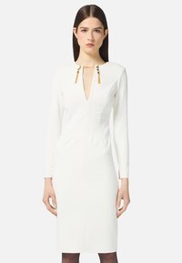 Frau in einem figurbetonten weißen Longsleeve-Kleid mit tiefem V-Ausschnitt und goldenen Quastendetails am Kragen vor einem schlichten Hintergrund.