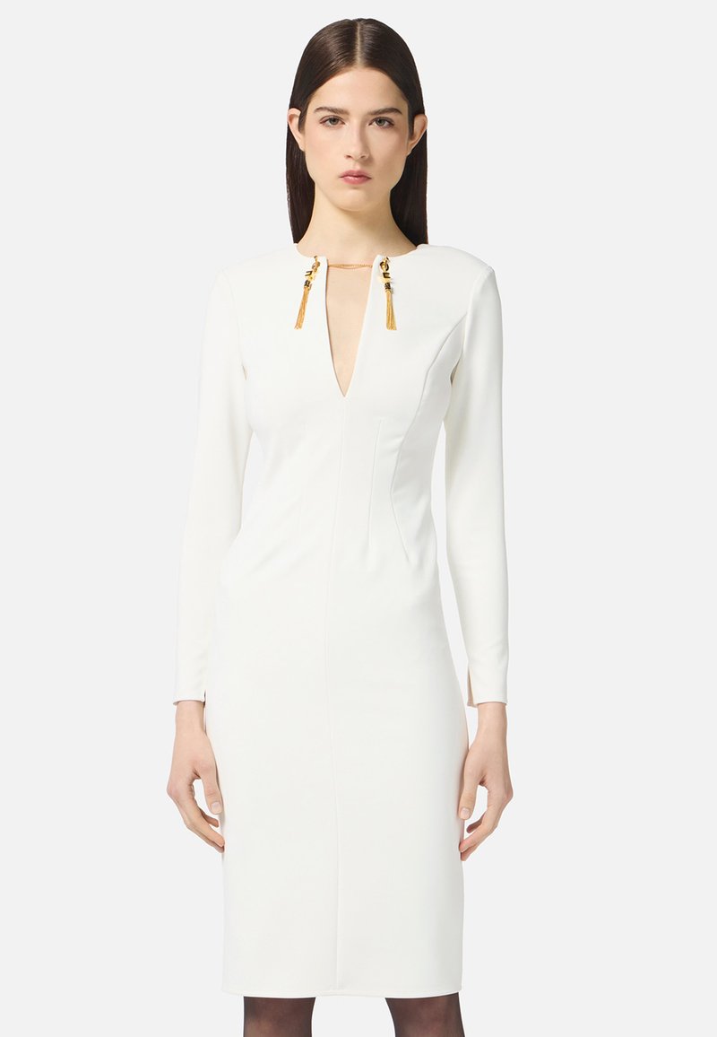 Frau in einem figurbetonten weißen Longsleeve-Kleid mit tiefem V-Ausschnitt und goldenen Quastendetails am Kragen vor einem schlichten Hintergrund.