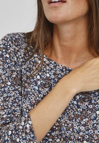 Haut à motif floral avec un col rond en bleu marine, rehaussé de touches blanches et brunes. Tissu doux et manches trois-quarts visibles.