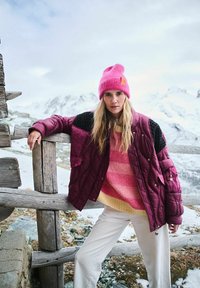 Bordová prešívaná bunda s čiernymi ramennými panelmi, spárovaná s pruhovaným svetrom v ružovej a žltej farbe a bielymi nohavicami. Na hlave jasne ružová čiapka.