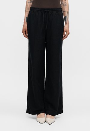 Personne portant un pantalon noir ample avec taille à cordon et des chaussures blanches à bout pointu, debout devant un fond uni.