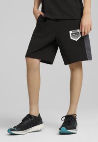 Shorts sportivi neri con un pannello grigio laterale, caratterizzati da un logo Puma bianco sulla coscia sinistra. Indossati con scarpe sportive nere.